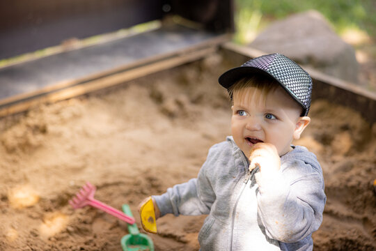Young child playing in sandbox wearing gray jacket and black cap, holding a toy shovel, with colorful sand toys visible in the background