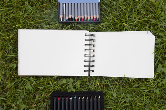 Top view of open blank spiral notebooks on green grass background, surrounded by sketching markers, drawing pencils and artist supplies.