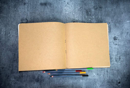 Top view of open kraft paper notebook with surrounded by pencils on a grey stone background.