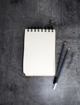Top view of open spiral A6 sketchbooks with surrounded by pencils on a grey stone background.