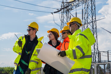 Fototapeta premium Four utility workers wear yellow hard hats and reflective vests. One holds a large paper blueprint, others gesture toward it. Tall metal power line tower stands behind them