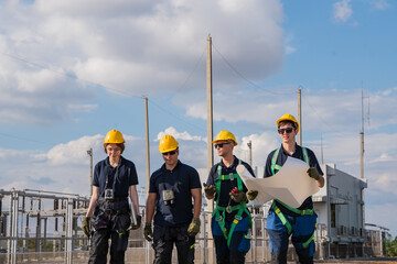 Fototapeta premium Four workers in hardhats and harnesses walk through a power substation. They hold a blueprint and discuss infrastructure plans. The diverse team focuses on renewable energy industry growth