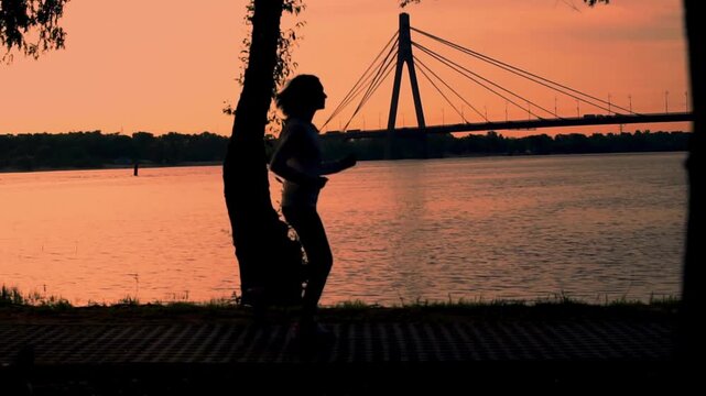 Silhouette of a runner jogging along the riverbank at sunset with a bridge in the background