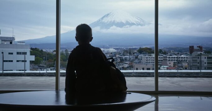 Cinematic silhouette of little boy staring out large window at distant Mt. Fuji with clouds - medium shot