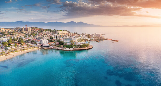 Kusadasi resort town on the Aegean Sea coastline, featuring a sandy beach, buildings, and clear turquoise water during sunset