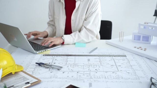 Architect working on new residential project using laptop, reviewing blueprints and drafting floor plans for modern building design with a 3d model on office desk