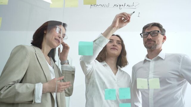 Business professionals collaborating at office meeting, developing strategy and brainstorming ideas on a transparent glass board, working together for project success and innovation
