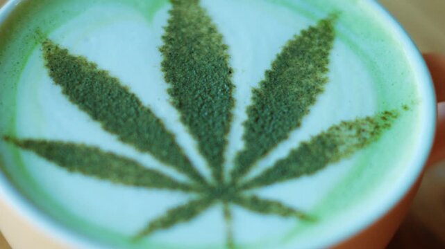 Top view of a matcha latte in a ceramic cup, decorated with powdered green tea forming a leaf pattern resembling a cannabis leaf