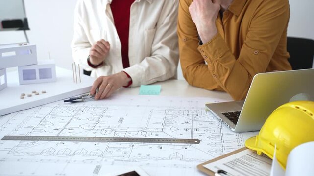 Engineering team collaborating on architectural blueprints, reviewing construction plans, and discussing building design concepts with tools and safety hard hat on table