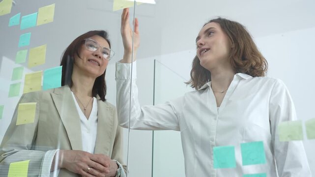 Diverse businesswomen collaborating and brainstorming innovative ideas while sticking and arranging colorful post it notes on a clear glass wall in a modern office meeting or workshop