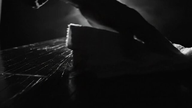 Low angle black and white shot of hand cleaning a worn wooden surface beneath a glowing light