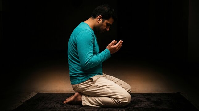 Devout Muslim man kneeling on a prayer mat and making dua with hands raised in a dark, quiet room