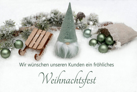Weihnachtliche Dekoration mit gr&uuml;nem Wichtel, Holzschlitten, Tannenzweigen und gr&uuml;nen Weihnachtskugeln. Im Schnee steht der Text Wir w&uuml;nschen unseren Kunden ein fr&ouml;hliches Weihnachtsfest'