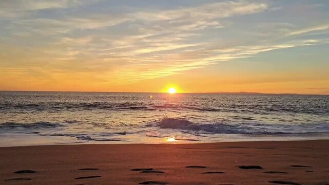 Golden sun dips below the horizon casting warm orange and yellow hues across the sky and ocean waves gently lapping the sandy shore with footprints
