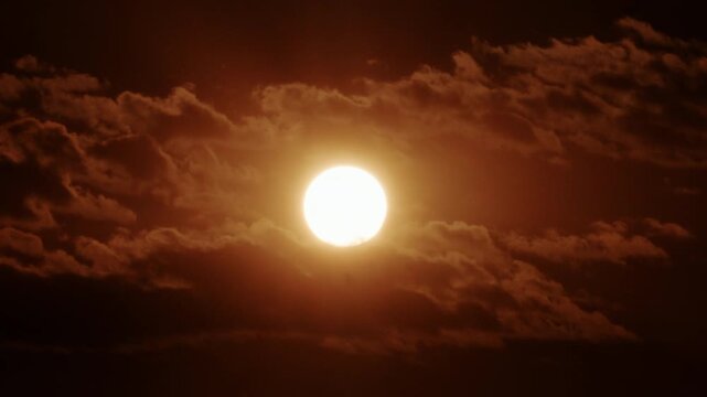 Close up of a bright sun partially covered by thick moving clouds, glowing in intense golden orange sunset tones