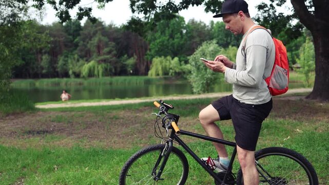 Man rider texts on smartphone sitting on bicycle at summer countryside. Sportsman surfs internet on cellphone resting during bike training
