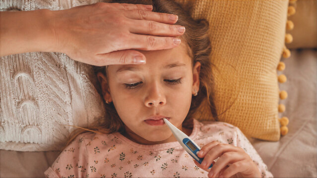 Sick Little Girl Lying in Bed with Digital Thermometer in Mouth while Mother Checks Her Temperature with Hand on Forehead