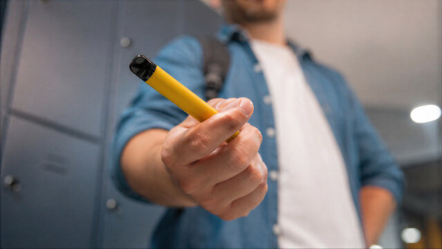 Young man holding a disposable vape pen in a school locker room hallway