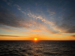 Obraz premium The glowing sun dips toward the horizon over gentle sea ripples. A warm orange and golden sky with soft clouds creates a peaceful, serene evening atmosphere.