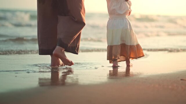 Close up of Asian parent and toddler girl feet walking barefoot on sandy beach at sunset golden hour. Hazy cinematic shot of family summer vacation and slow living bonding. Ideal for travel, parenting