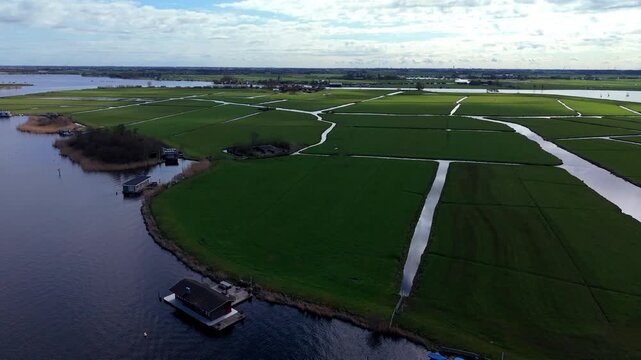 An aerial drone shot revealing a picturesque Dutch polder landscape with vibrant green fields interlaced with drainage canals, bordered by a large lake featuring houseboats under a cloudy sky.