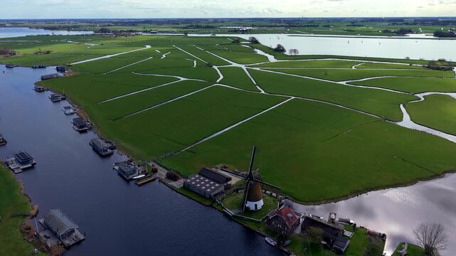 Aerial drone view of a traditional Dutch windmill standing along a canal with houseboats, set against a vast green polder landscape under a partly cloudy sky.