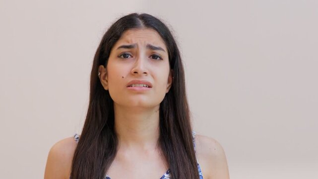 Stressed young female showing worry  nervousness  or unease about something. Closeup shot of a troubled Indian girl feeling anxious while standing against a wall background - facial expressions con...