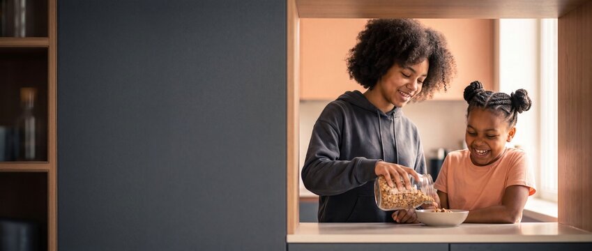 Black sisters preparing morning breakfast eating cereal together in modern kitchen. Happy African American family, siblings laughing, healthy lifestyle, wellness, copy space panoramic background.