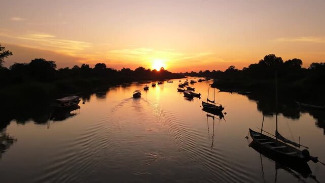 Serene river landscape at sunset with numerous boats of various sizes docked and moving on the calm water, silhouetted trees lining the banks under a vibrant orange and yellow sky reflecting on the