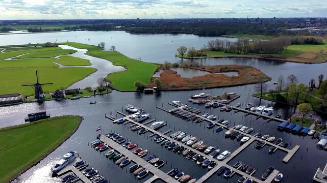 An aerial drone shot looking over a large marina filled with docked boats, expanding into a green Dutch polder landscape with a traditional windmill and lakes.