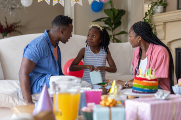 Obraz premium African American family celebrating in living room; girl in tiara opening gift near rainbow cake