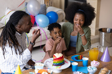 Obraz premium African American family eating rainbow cake, sitting at living-room table with blue cups