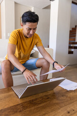 Fototapeta premium Asian man sitting in living room wearing mustard tee and denim shorts reviewing papers with laptop