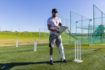 Obraz premium Man adjusting batting pad while standing on turf at cricket nets with stumps nearby