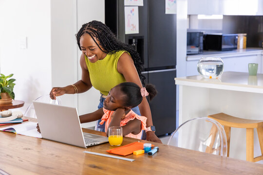African American mother leaning over guiding child at kitchen table using laptop and markers