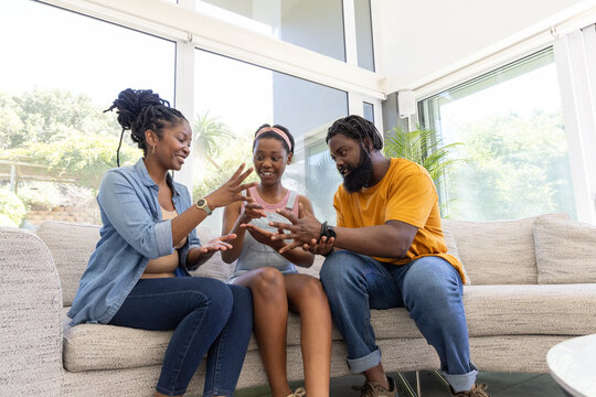 African family playing hand game on beige sofa by large windows, wearing denim and mustard shirts