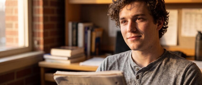 Male student studying at desk looking out window in collegiate library. Young man reading notes with books and warm sunlight background, visualizing success, higher education in college dorm room.