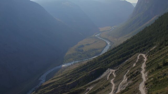 Drone footage of the winding dirt road on Katu-Yaryk Pass overlooking the Chulyshman River and deep mountain valley in Altai Republic, Russia, in soft morning haze