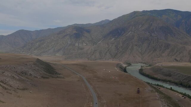 Drone footage of Chuysky Trakt road winding through a wide mountain valley beside a turquoise river, Altai Republic, Russia, dramatic ridges and cloudy sky