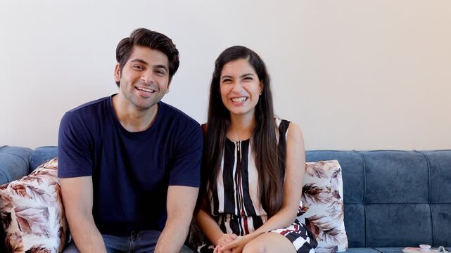 Indian boyfriend-girlfriend cheerfully greeting their relatives on a video call. Medium shot of a young couple happily talking to their friends while sitting together on a couch - lifestyle concept