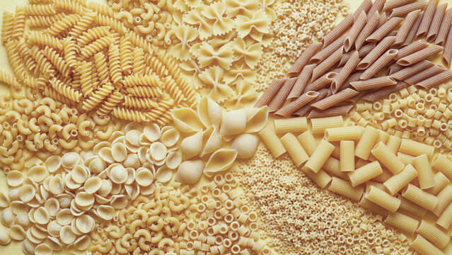 A flat lay of assorted raw pasta shapes including fusilli, penne, farfalle, and shells densely arranged in a creative, textured pattern on a pale yellow background.