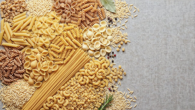 Assorted raw pasta shapes like penne, fusilli, spaghetti, and macaroni arranged on the left of a textured grey linen background, leaving ample copy space on the right for design composites.