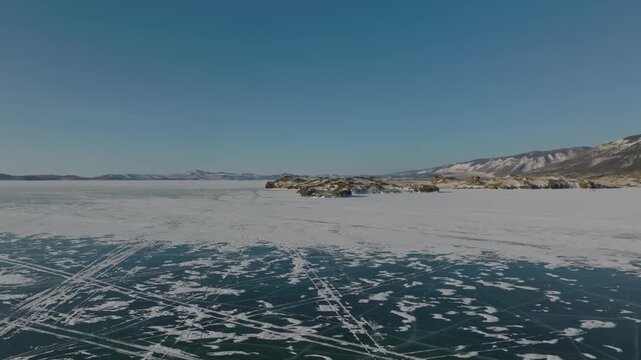 Drone footage of a vast frozen surface of Lake Baikal with rocky islets, ice patterns, and snow-covered coast under clear sky, Siberia, Russia