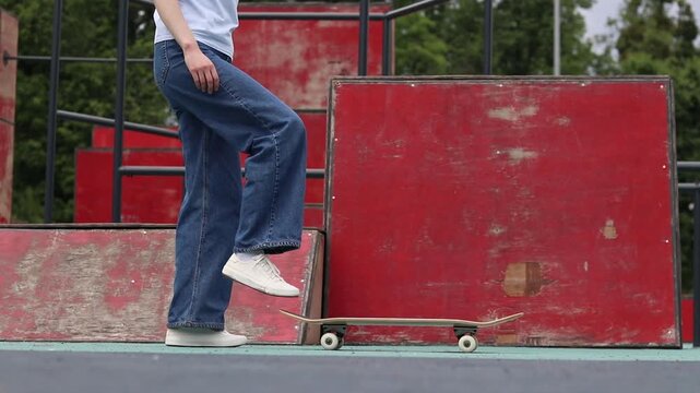 Girl in wide jeans getting ready to skateboard in skate park. Skater holding board in front of red ramp. Young woman doing extreme sports outdoors. Urban style and street sports