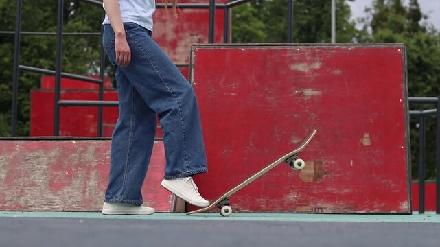 Girl in wide jeans getting ready to skateboard in skate park. Skater holding board in front of red ramp. Young woman doing extreme sports outdoors. Urban style and street sports