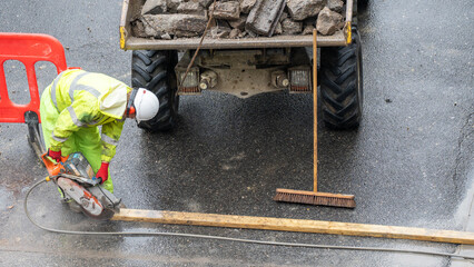 Construction worker in yellow vest cutting wet road with saw near parked dumper truck © Preethiloki
