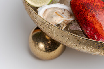 Detail of lobster claw and oyster beside golden bowl rim. Overhead close up of red shell, open bivalve and lime with condensation on textured brass edge. © Alisa