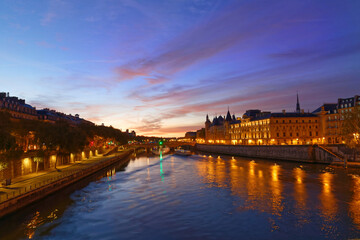 Obraz premium Breathtaking blue-hour panorama of the River Seine in Paris, with the illuminated Conciergerie and its medieval towers glowing in golden light on the right bank.