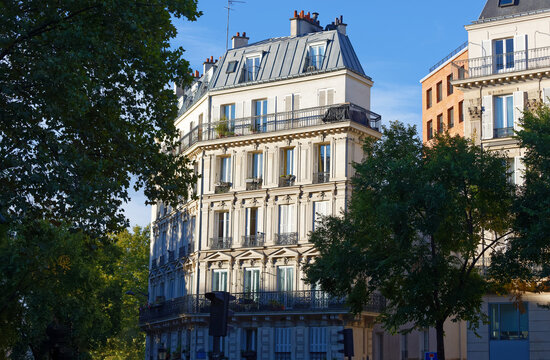 Classic Haussmann-style apartment building on Boulevard de Magenta, Paris, framed by lush autumn trees under a vivid blue sky. The cream limestone facade features elegant wrought-iron balconies,