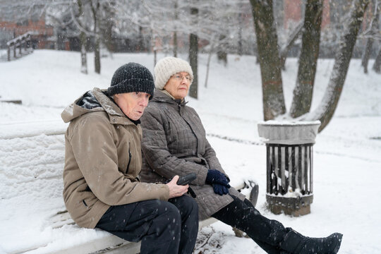 Elderly couple sitting close together on bench in snowy winter park. Senior pair sharing quiet tender moment on cold peaceful day. Caring companions resting side by side with frozen lake view